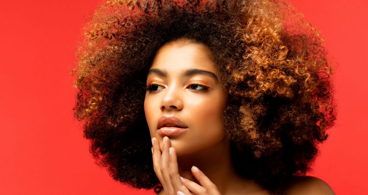 Mulher com a mão no queixo, usando maquiagem alaranjada nos olhos com cabelo cacheado em tom ruivo doce de leite.